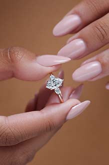 A woman with shell-pink nails slips a custom lab created diamond ring set with side stones onto a yellow gold band, onto her finger.