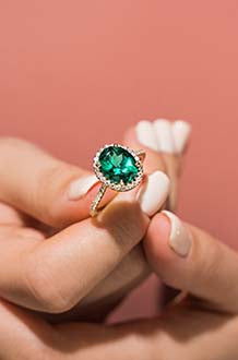 A woman holds a MiaDonna ring featuring a rich emerald green colored, oval shaped gemstone with a halo and band of lab created diamonds.